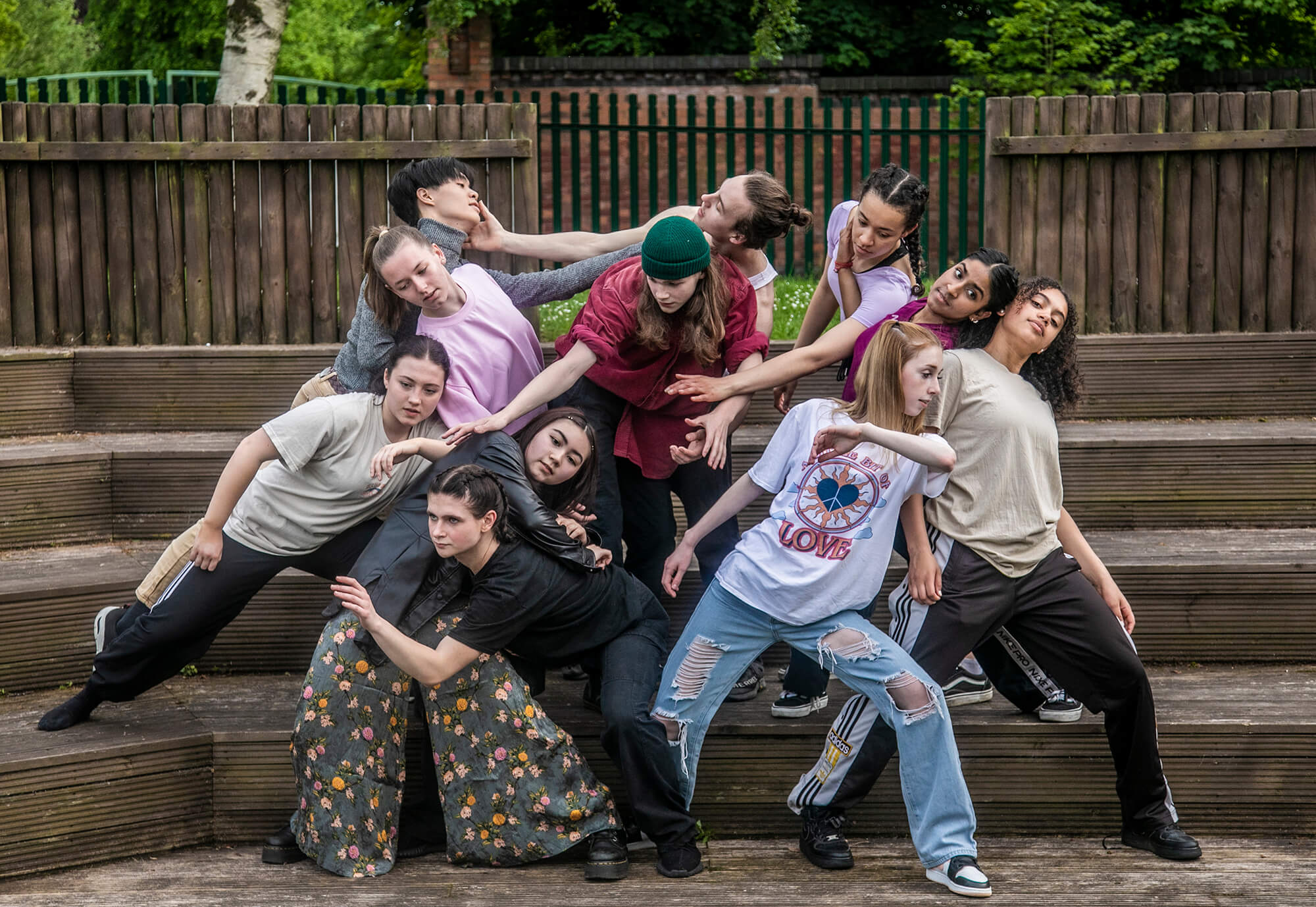 Eleven young male and female dancers huddled in a group on wooden steps moving around eachother with stretched arms. Causually dressed. 