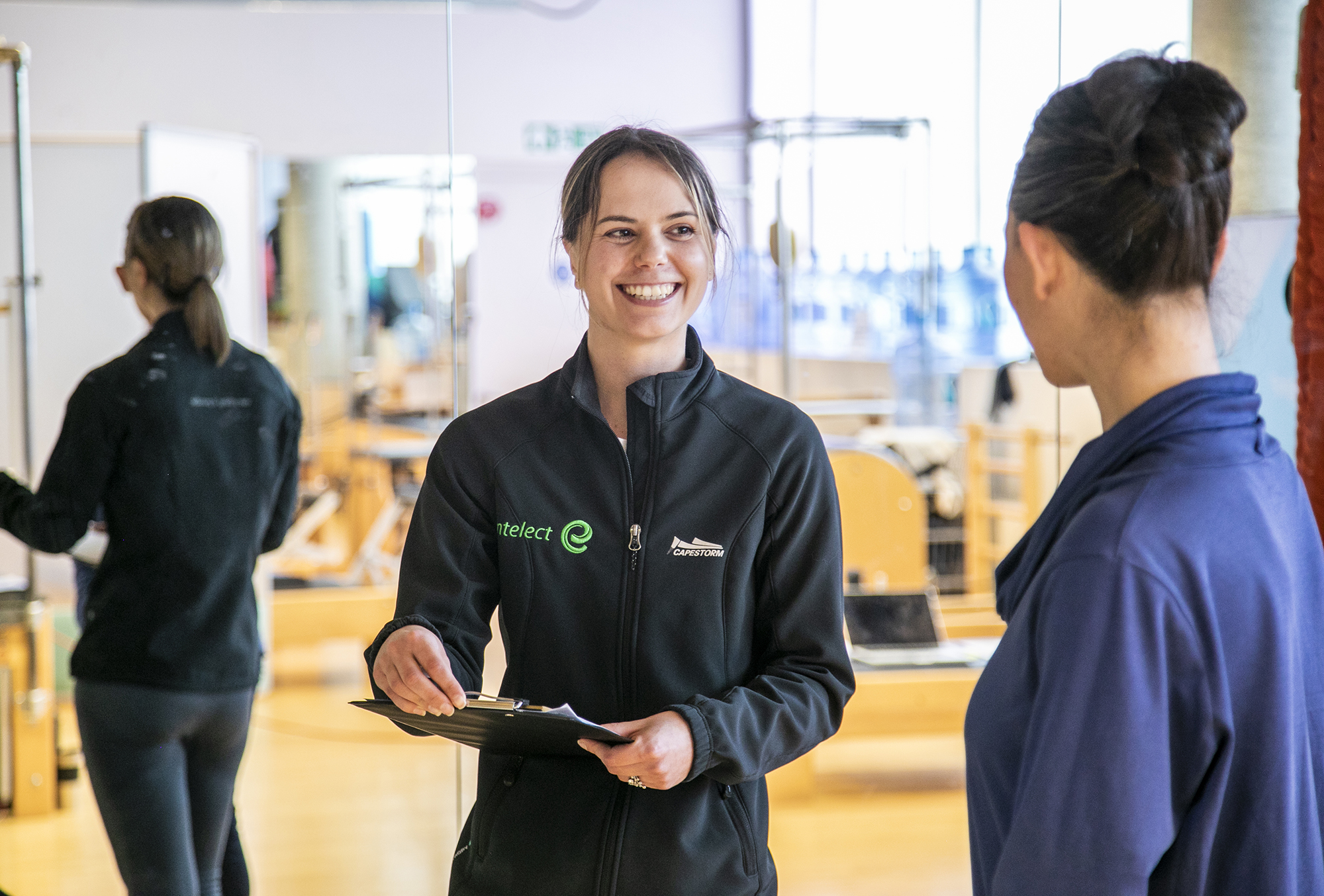 White female with brown tied back hair wearing black zip up jumper holding a clipboard and smiling whilst looking at a white female with tied up black hair wearing purple jumper whos face is turned away from the camera. With mirrored background in a fitness setting. White female with brown tied back hair wearing black zip up jumper holding a clipboard and smiling whilst looking at a white female with tied up black hair wearing purple jumper whos face is turned away from the camera. With mirrored background in a fitness setting.