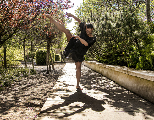 Global majority male dancer wearing black dress with short black hair. One leg kicked high with arms following the line. On a path between trees with lots of tree shadows.  