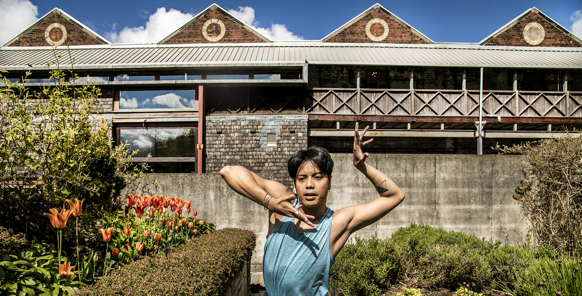 Global majority male dancer with short black hair, centre of the image with hand framing his face. Wearing blue tank top in front of tulips and triangular shaped building  Global majority male dancer with short black hair, centre of the image with hand framing his face. Wearing blue tank top in front of tulips and triangular shaped building