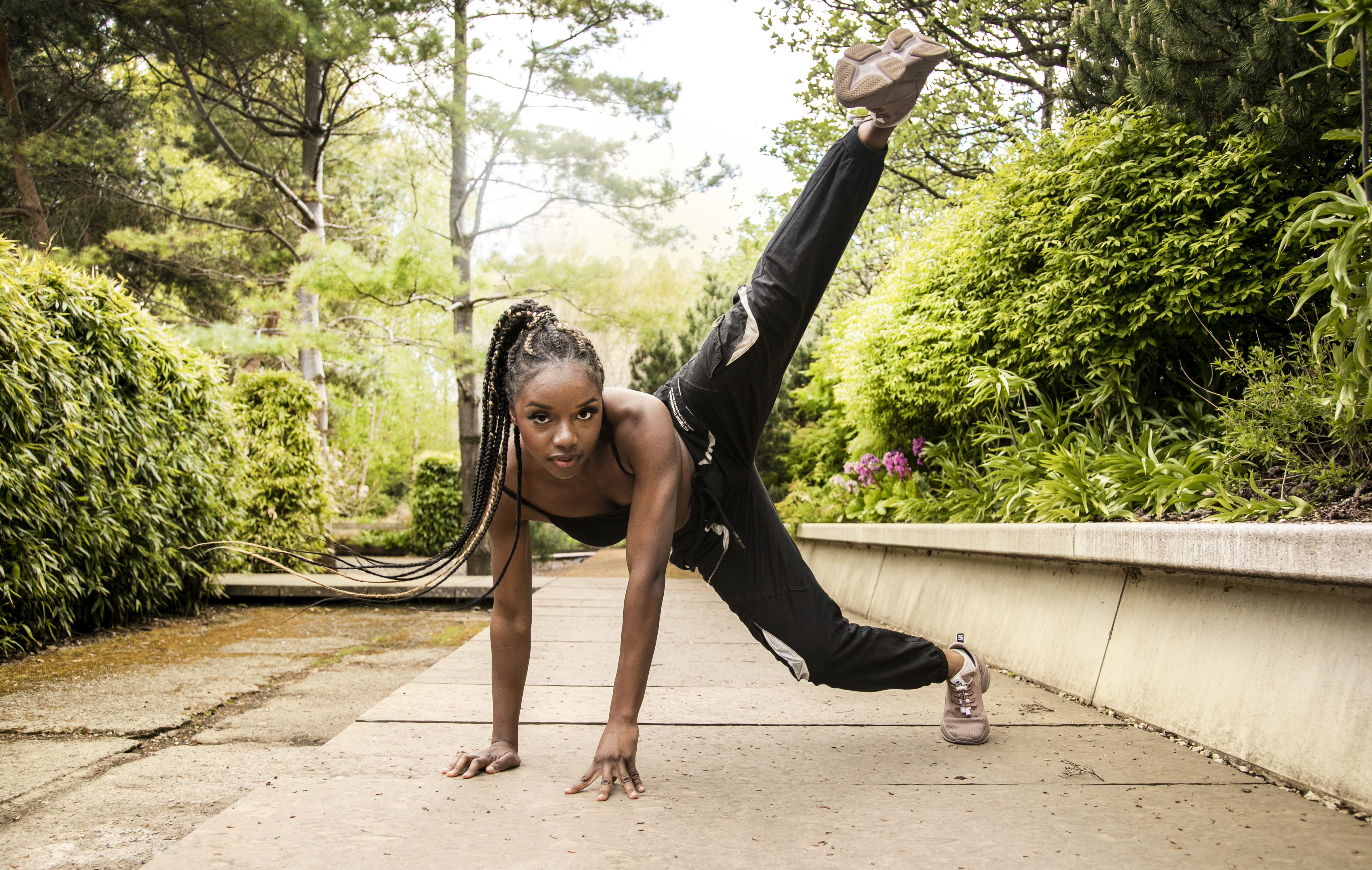 Global majority female hands on the floor kicking leg in the air with braided hair flicking to the left. Wearing black trousers and strap top. In a garden on a path Global majority female hands on the floor kicking leg in the air with braided hair flicking to the left. Wearing black trousers and strap top. In a garden on a path