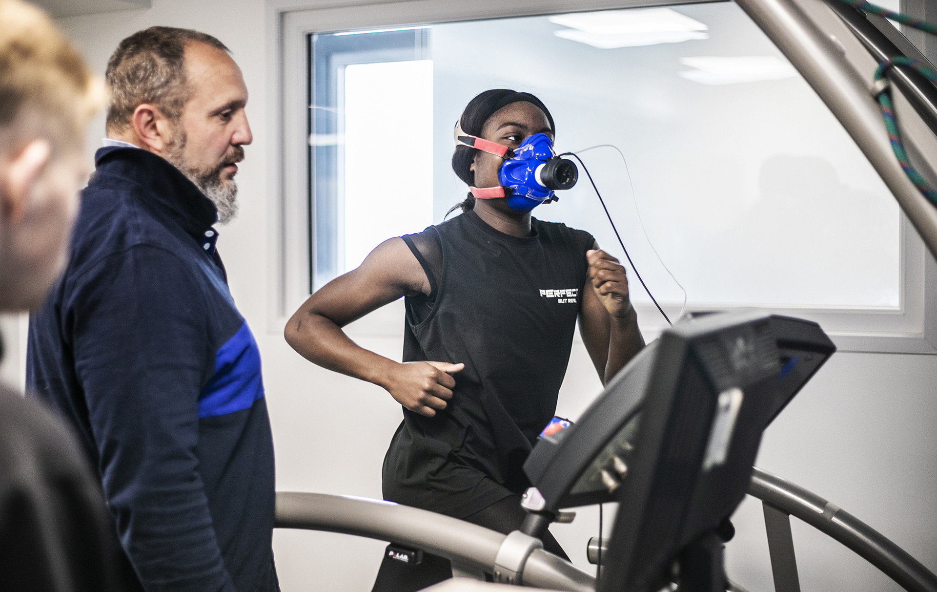 Global majority female dancer running on a treadmill wearing face-mask to measure cardiovascular fitness. White male physiologist looking over to observe stats on the machine. In a white laboratory setting with a window in the background Global majority female dancer running on a treadmill wearing face-mask to measure cardiovascular fitness. White male physiologist looking over to observe stats on the machine. In a white laboratory setting with a window in the background