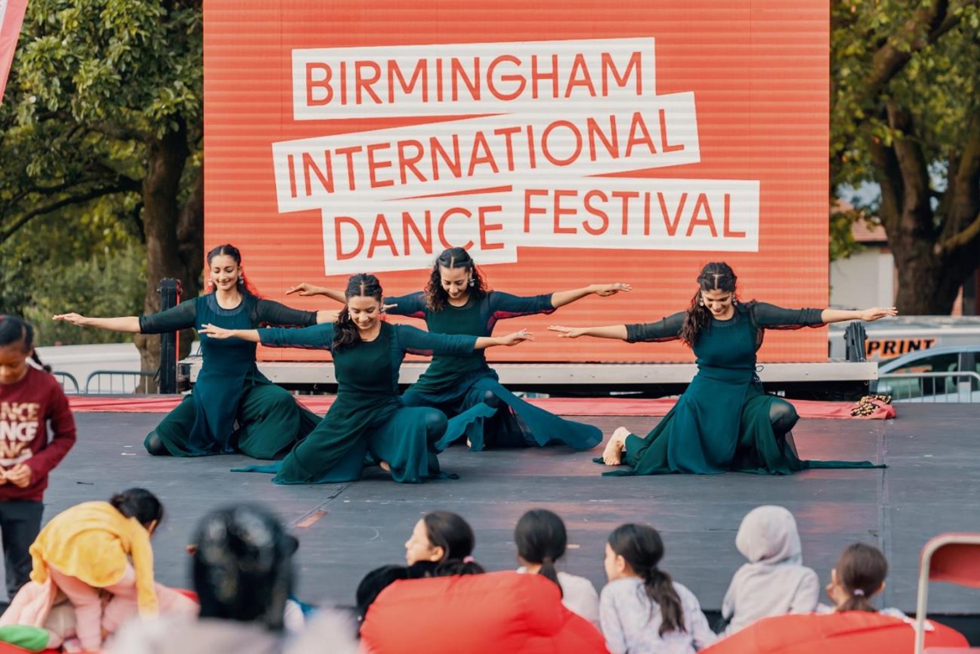 four South Asian dancers all female on one knee with arms out wide in front of large red sign saying Birmingham international dance festival. In front of an outdoor audience. four South Asian dancers all female on one knee with arms out wide in front of large red sign saying Birmingham international dance festival. In front of an outdoor audience.