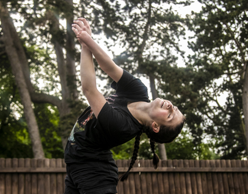 White female dancer with limb difference arms pointing to the  top left hand side of the image with arched back. Wearing black t-shirt and two long plaits. In front of soft focus trees