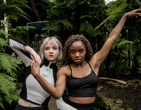 Two dancers arms intertwined looking at the camera in a greehouse with plants. One white femles with blonde hair black and white long sleve top. One black female with long braided hair in black tank top. 