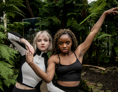 Two dancers arms intertwined looking at the camera in a greehouse with plants. One white femles with blonde hair black and white long sleve top. One black female with long braided hair in black tank top. Two dancers arms intertwined looking at the camera in a greehouse with plants. One white femles with blonde hair black and white long sleve top. One black female with long braided hair in black tank top.