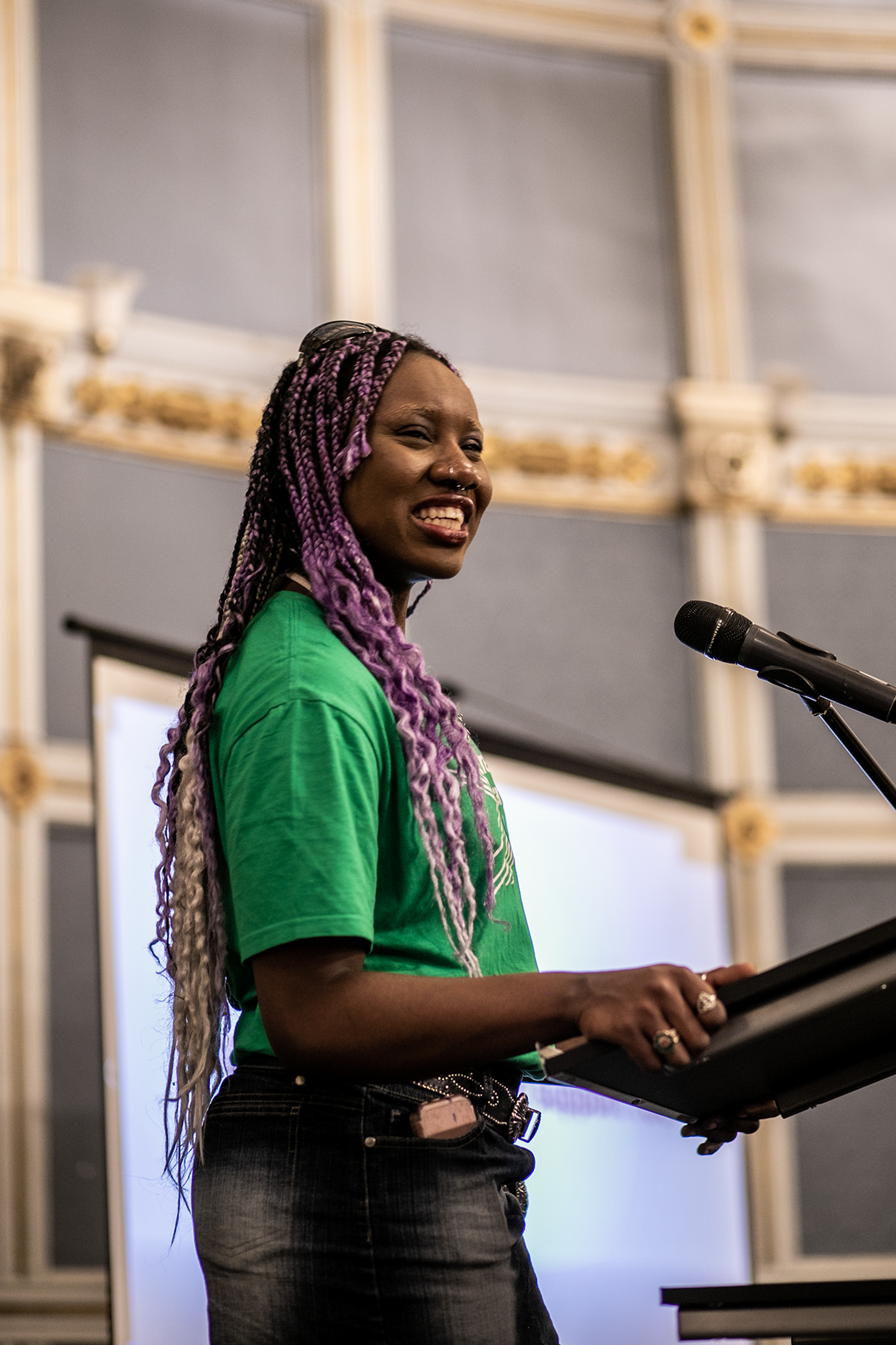 Black non binary person with purple braided hair Dance ambassador talking on stage at One Dance UK AGM wearing green tshirt 