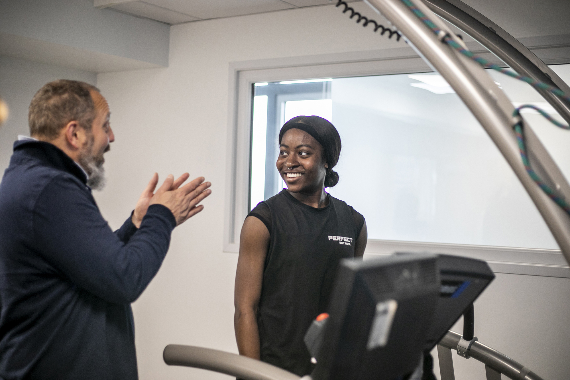 Global majority female dancer standing on a treadmill smiling whilst white male physiologist is explaining something to her. In a white laboratory setting with a window in the background Global majority female dancer standing on a treadmill smiling whilst white male physiologist is explaining something to her. In a white laboratory setting with a window in the background