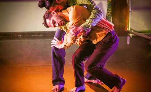 two male dancers huddled over each other wearing bright clothes with an orange spotlight