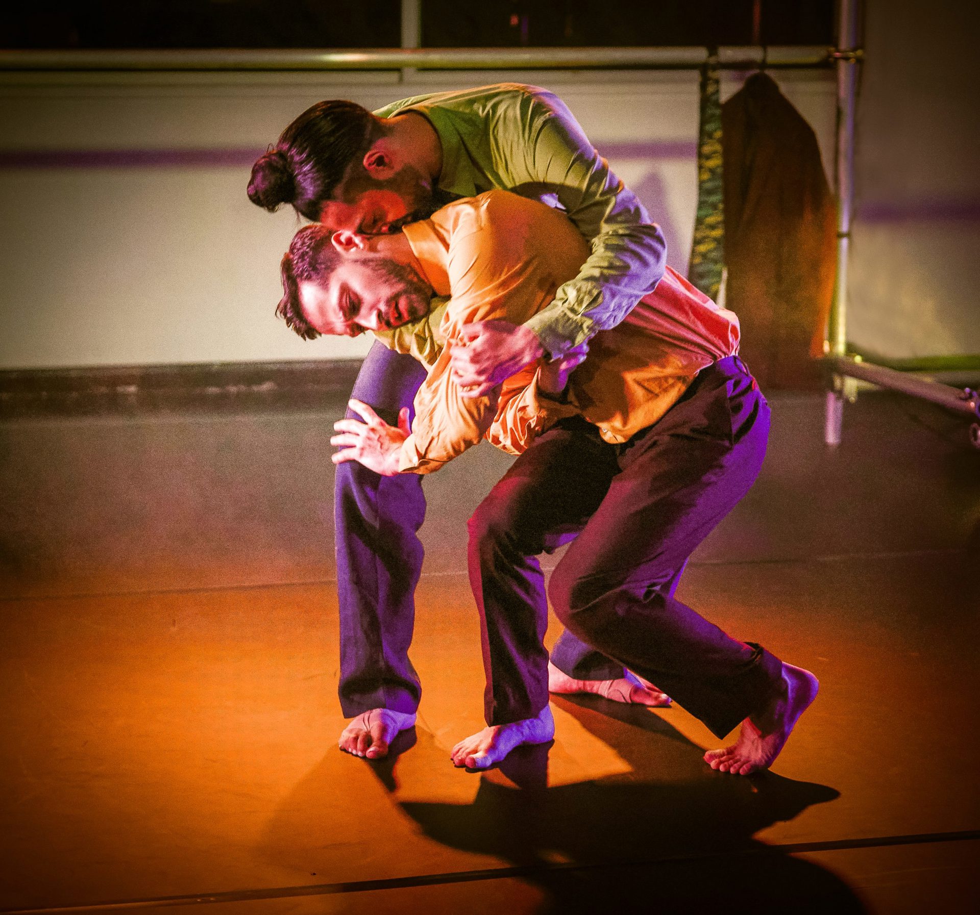 two male dancers huddled over each other wearing bright clothes with an orange spotlight