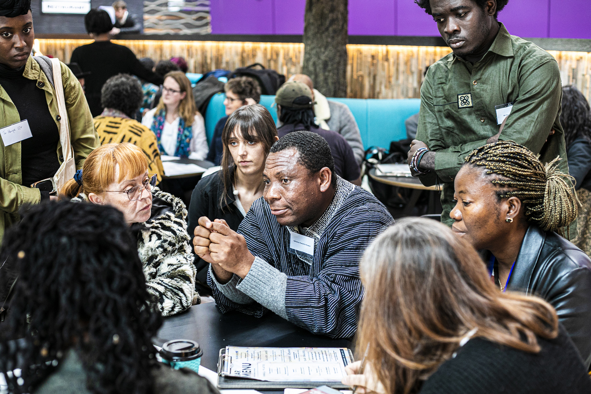 Global majority male in the centre of the image sitting at a table holding his hands together with a group of people listening to him of mixed ages, genders and ethnicities. Global majority male in the centre of the image sitting at a table holding his hands together with a group of people listening to him of mixed ages, genders and ethnicities.