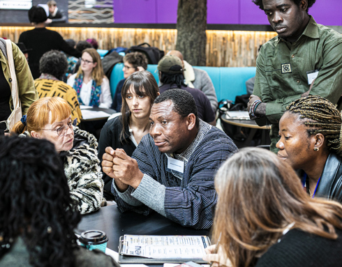 Global majority male in the centre of the image sitting at a table holding his hands together with a group of people listening to him of mixed ages, genders and ethnicities. Global majority male in the centre of the image sitting at a table holding his hands together with a group of people listening to him of mixed ages, genders and ethnicities.