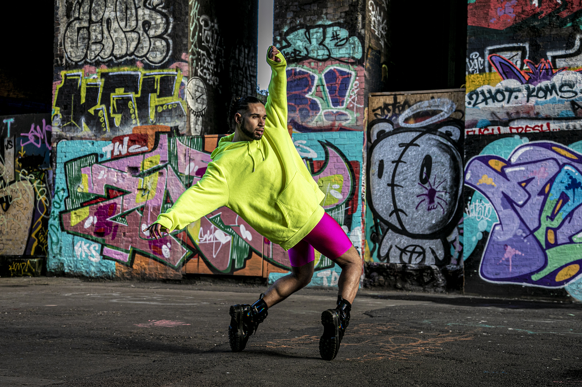 LGBTQ+ global majority male dancer with short dreads wearing neon yellow jumper with pink leotard shorts and black boots. Hands around the face, standing top toes with bent knees leaning back with arms out stretched. In front of colourful graffiti wall. LGBTQ+ global majority male dancer with short dreads wearing neon yellow jumper with pink leotard shorts and black boots. Hands around the face, standing top toes with bent knees leaning back with arms out stretched. In front of colourful graffiti wall.