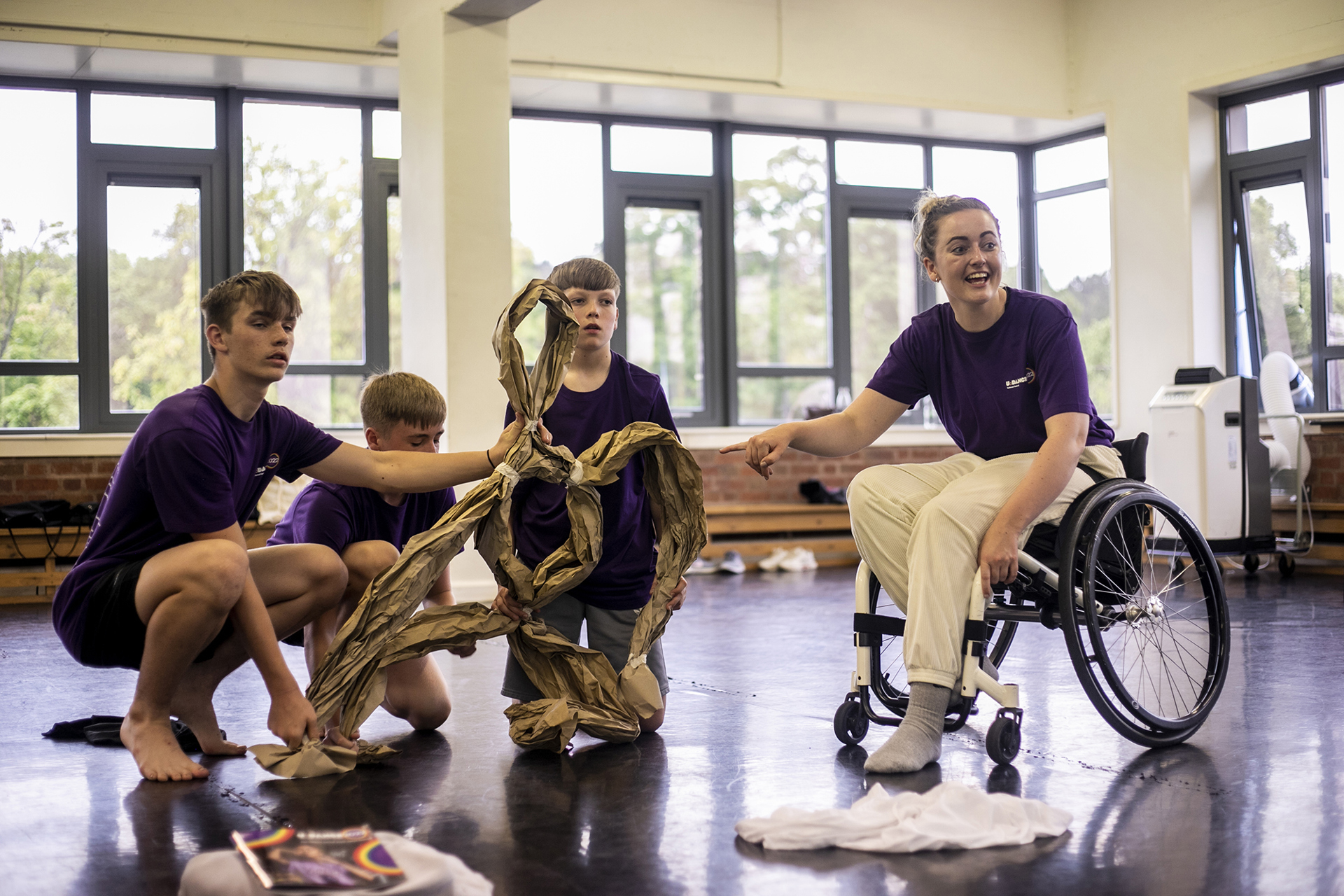 three young white boys holding up a paper puppet with a white female wheel chair dancer holding the puppets arms and smiling, in a dance studio. three young white boys holding up a paper puppet with a white female wheel chair dancer holding the puppets arms and smiling, in a dance studio.