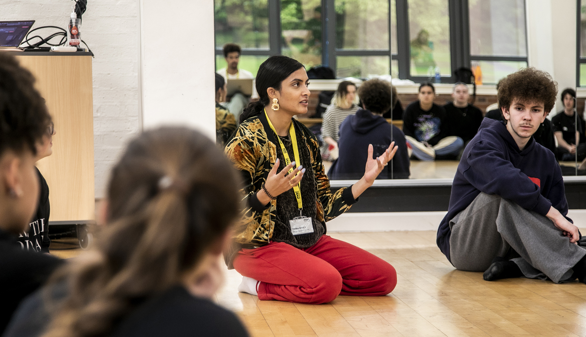 golbal majority dance teacher sitting on her knees speaking to a room full of dance students gesturing her hands, with mirror reflecting the students behind her. Wearing red trousers and colourful jacket. 