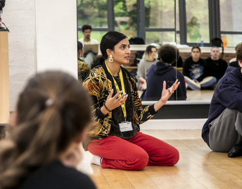 golbal majority dance teacher sitting on her knees speaking to a room full of dance students gesturing her hands, with mirror reflecting the students behind her. Wearing red trousers and colourful jacket. golbal majority dance teacher sitting on her knees speaking to a room full of dance students gesturing her hands, with mirror reflecting the students behind her. Wearing red trousers and colourful jacket.