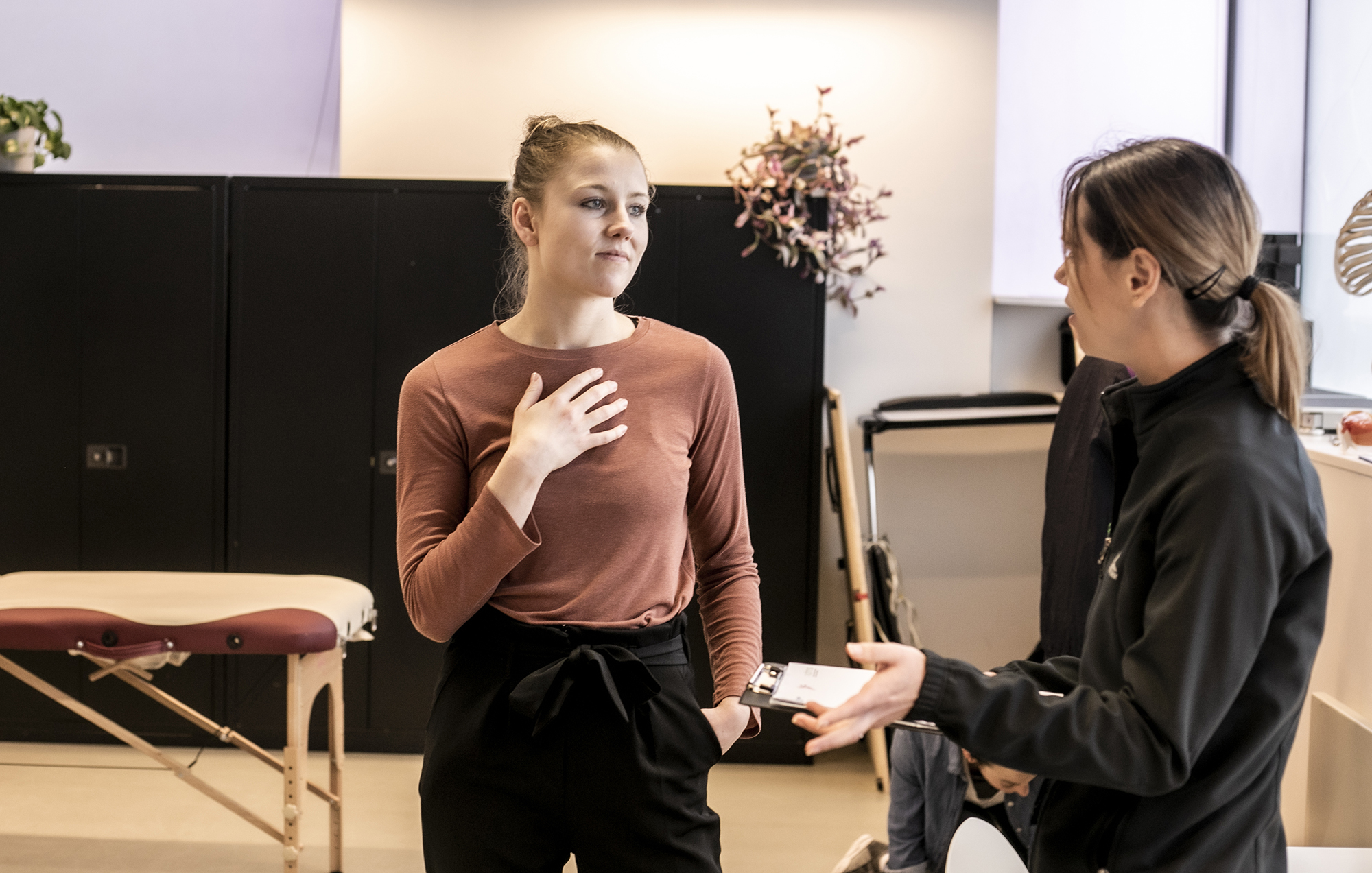 White female healthcare practitioner holding clipboard explainig something to white female dancer who is holding hand to heart in a healthcare environment with physio table in the background.