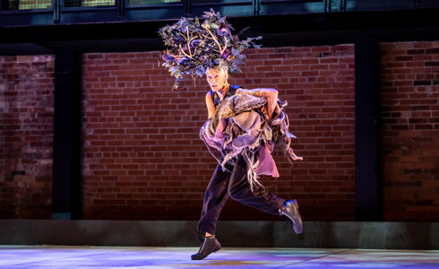 white female dancer running across the stage with large tree head piece carrying lots of clothes with brick background