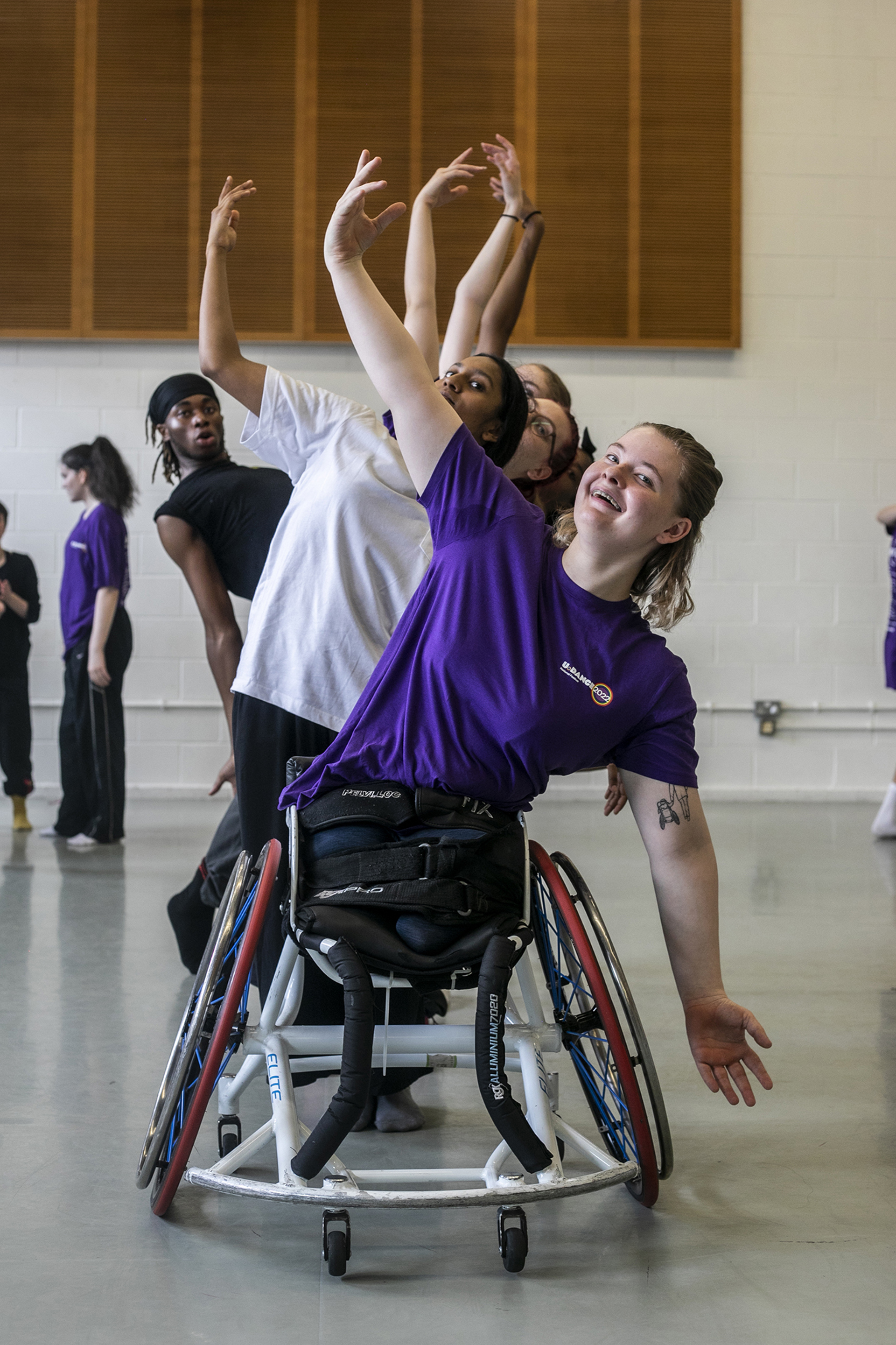 White female dancer in a wheel chair lifting one arm above her head with a mix of male and female, different raced dancers standing behind doing the same lifting arm pose, in a dance studio White female dancer in a wheel chair lifting one arm above her head with a mix of male and female, different raced dancers standing behind doing the same lifting arm pose, in a dance studio