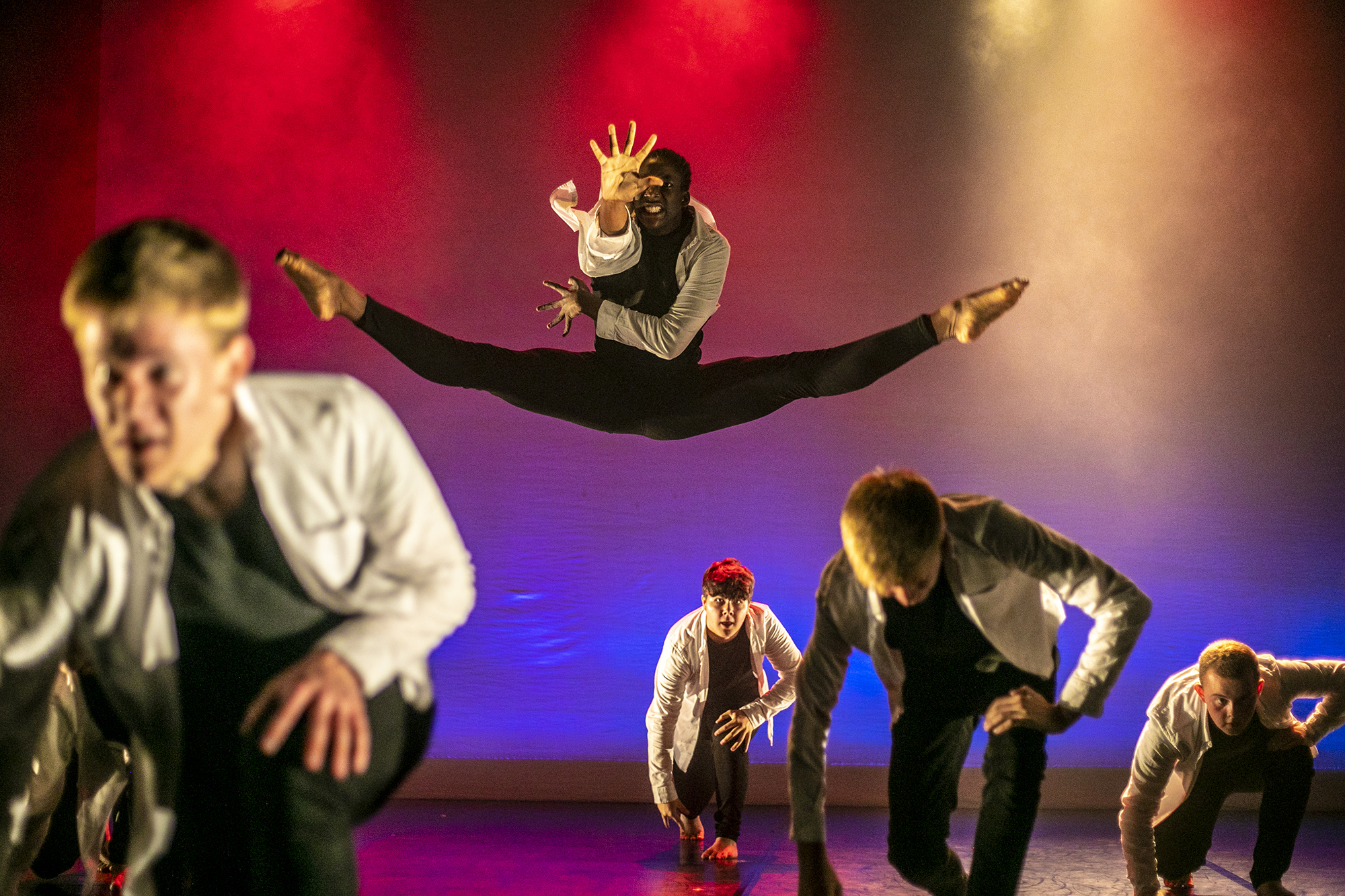 male dancers on U.dance stage wearing white shirts and black trousers. Four white young males crouched down one male black dancer jumpinging the box splits leaning forward with one hand stretched out. Lighting pink blue and red spotlights male dancers on U.dance stage wearing white shirts and black trousers. Four white young males crouched down one male black dancer jumpinging the box splits leaning forward with one hand stretched out. Lighting pink blue and red spotlights