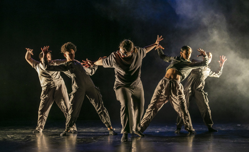 five female dancers one in the center with arms puhing out the other four leaning back from the push with arm out to the side. One a dark stage with smoke over back light on the right hand side. All wearing dark neutral tones