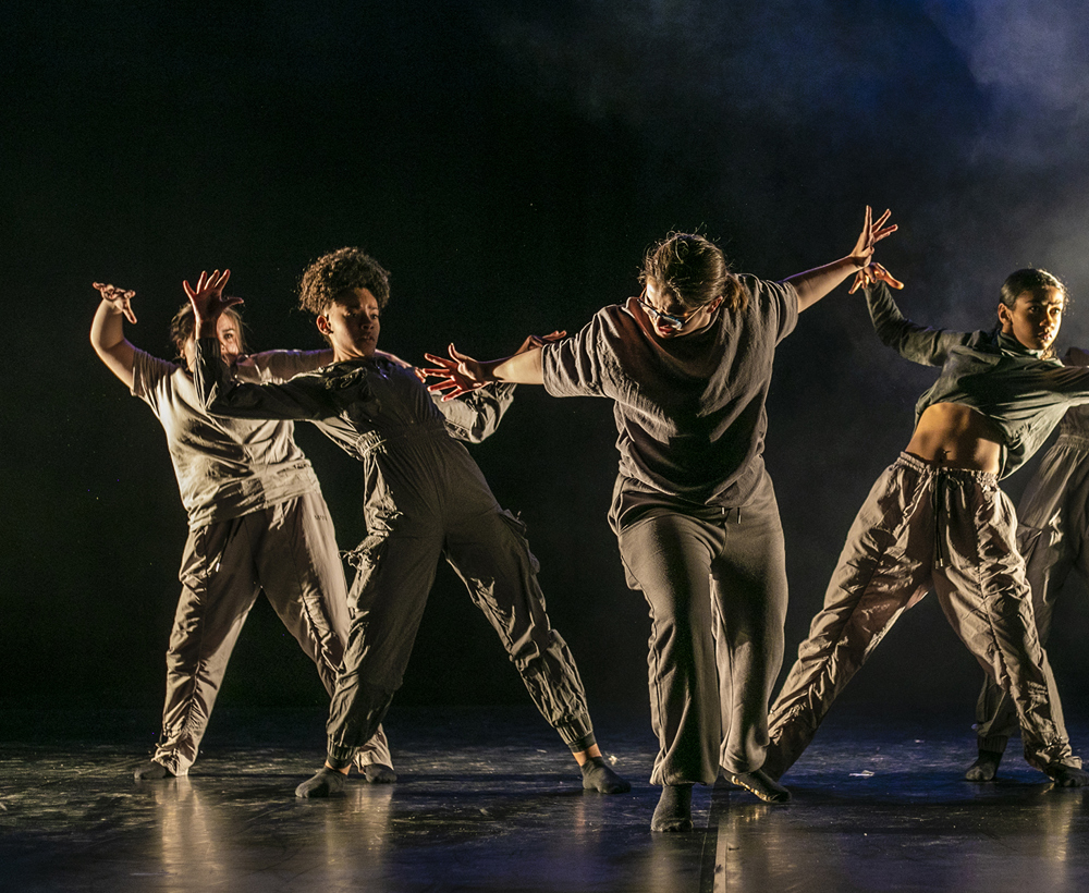 five female dancers one in the center with arms puhing out the other four leaning back from the push with arm out to the side. One a dark stage with smoke over back light on the right hand side. All wearing dark neutral tones
