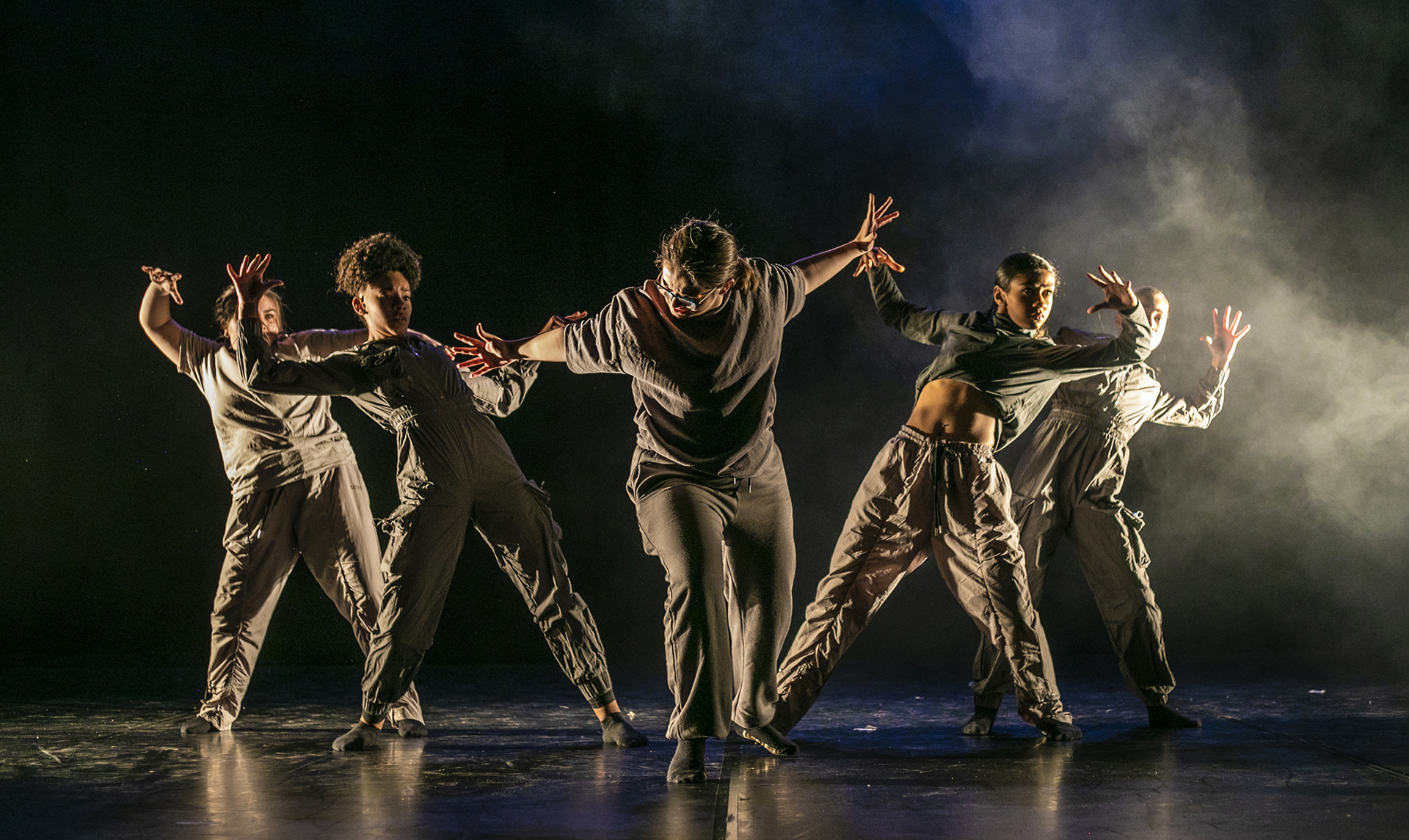 five female dancers one in the center with arms puhing out the other four leaning back from the push with arm out to the side. One a dark stage with smoke over back light on the right hand side. All wearing dark neutral tones