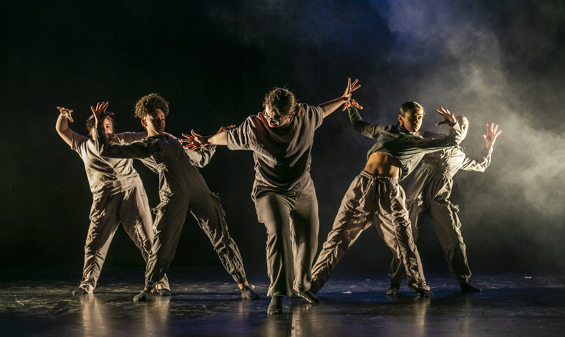 five female dancers one in the center with arms puhing out the other four leaning back from the push with arm out to the side. One a dark stage with smoke over back light on the right hand side. All wearing dark neutral tones five female dancers one in the center with arms puhing out the other four leaning back from the push with arm out to the side. One a dark stage with smoke over back light on the right hand side. All wearing dark neutral tones