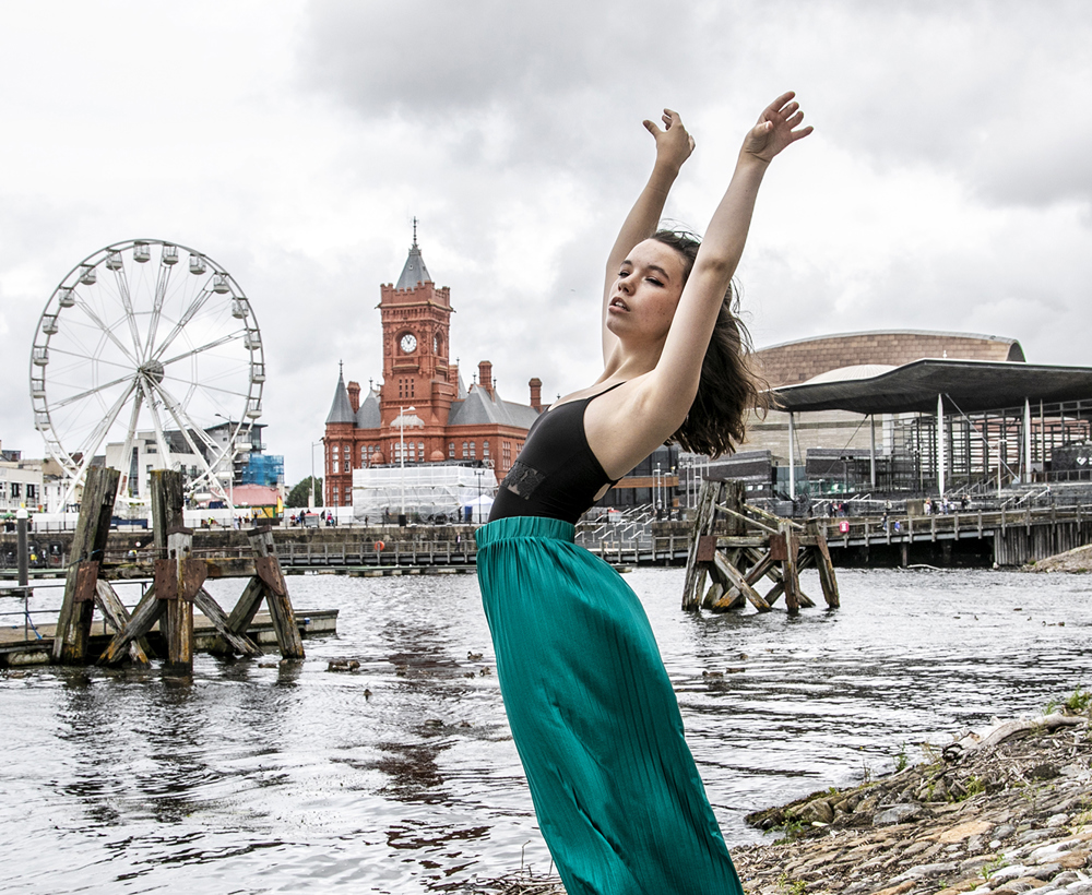 dancer with arched back and arms above head infront of Cardiff bay skyline. Young white female wearing black leotard and green silk trousers