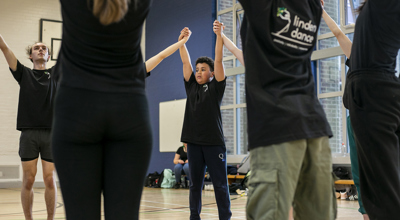 Dance circle with young people holding hands with the joint hands in the air. All wearing black PE kit in a sports hall.