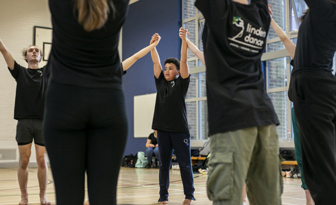 Dance circle with young people holding hands with the joint hands in the air. All wearing black PE kit in a sports hall.