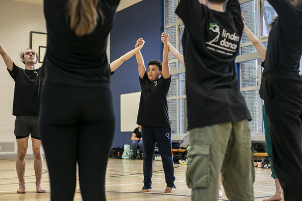 Dance circle with young people holding hands with the joint hands in the air. All wearing black PE kit in a sports hall.