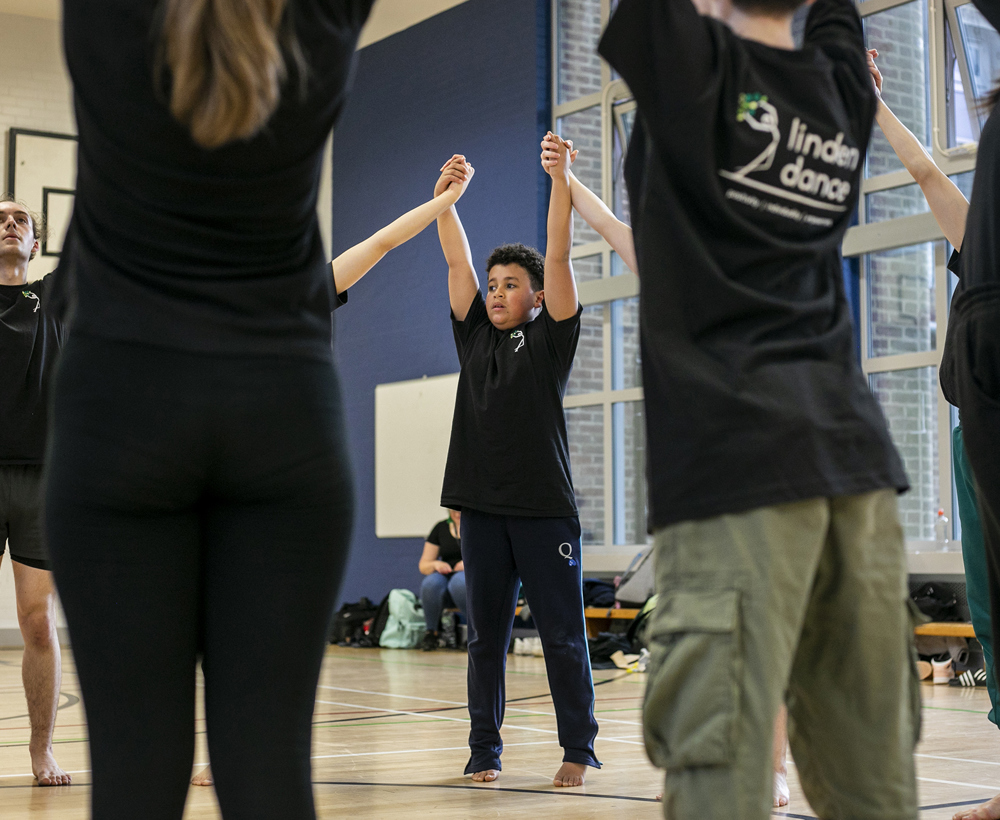 Dance circle with young people holding hands with the joint hands in the air. All wearing black PE kit in a sports hall.