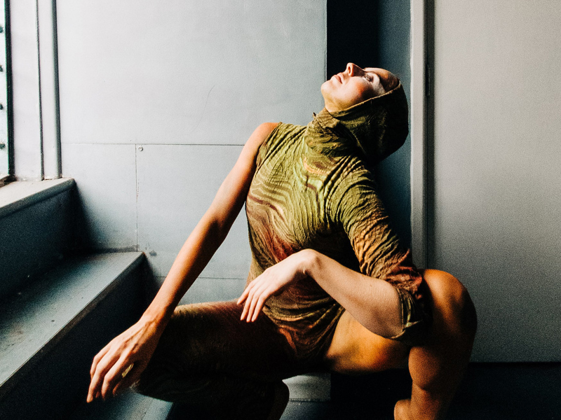 Image of Helen Calcutt. White female squatted down looking to the top left wearing green one sleeved leotard with a hood. Image of Helen Calcutt. White female squatted down looking to the top left wearing green one sleeved leotard with a hood.