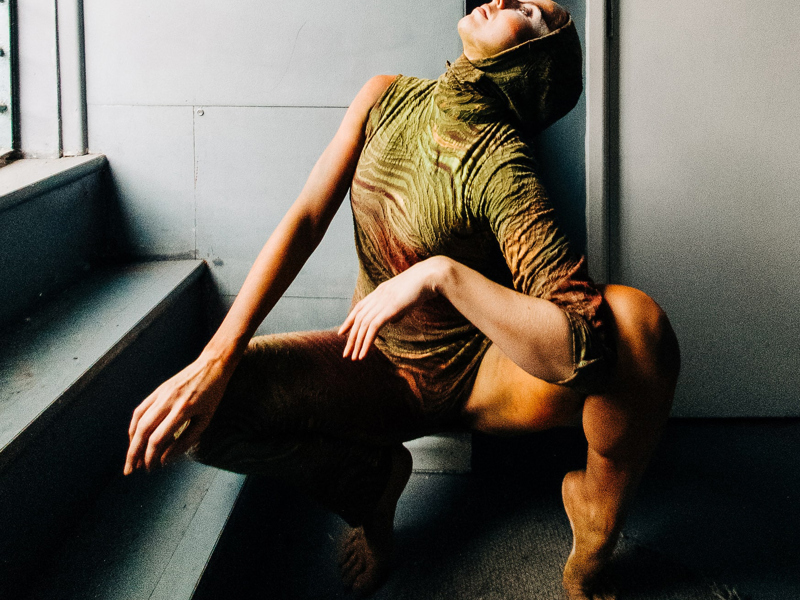 Image of Helen Calcutt. White female squatted down looking to the top left wearing green one sleeved leotard with a hood.  Image of Helen Calcutt. White female squatted down looking to the top left wearing green one sleeved leotard with a hood.