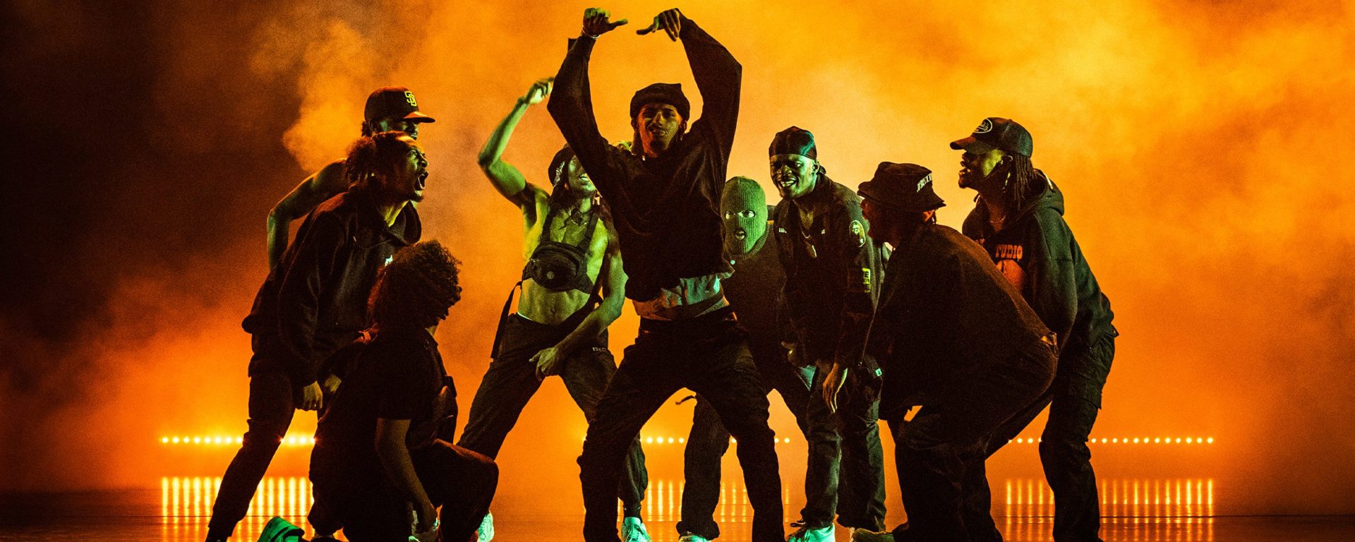Nine male dancers huddled around eachother shouting and cheering at central dancer who has his arms in the air. All wearing dark clothes on a dark stage with orange light over smoke Nine male dancers huddled around eachother shouting and cheering at central dancer who has his arms in the air. All wearing dark clothes on a dark stage with orange light over smoke